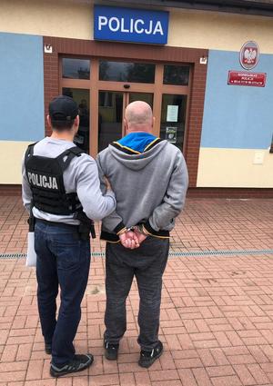 Teren przed KP w Białych Błotach. Policjant prowadzi zatrzymanego 46-latka.