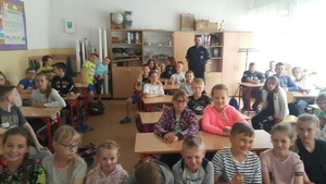 Zdjęcie podczas zajęć w klasie lekcyjnej. Uczniowie siedzą w ławkach. Wśród nich stoi policjant.