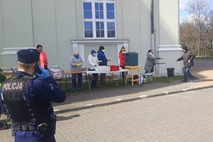 wolontariusze i pracownicy Bazyliki rozdają osobom posiłek i ulotki informujące o nowych obostrzeniach. Z przodu stoi policjant i rozmawia przez telefon