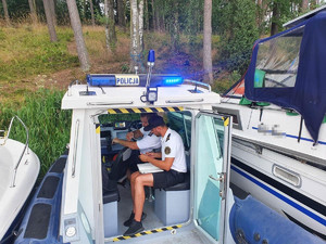 Policjanci wewnątrz policyjnej łodzi sporządzają dokumentację. Obok zacumowane inne łodzie.