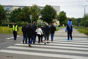 Policjant i &quot;polfinek&quot; pomagają grupie osób niepełnosprawnych pokonać przejście dla pieszych.