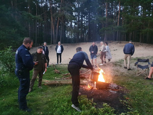 Policjant i grupa osób przy palącym się ognisku w lesie. Mężczyzna polewa wodą palące się ognisko.
