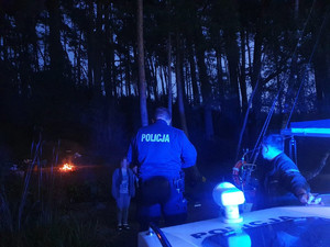 Po zmroku policjant na policyjnej łodzi motorowej ujawnił palące się ognisko w lesie i rozmawia z kobietą.