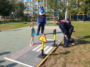 Policjant instruuje dziecko jak przechodzić przez przejście dla pieszych. Przygląda się temu policyjny Polfinek.