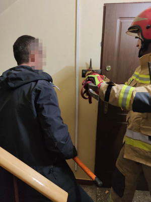 strażak wyłamuje zamek od drzwi. Obok stoi policjant w cywilnym ubraniu