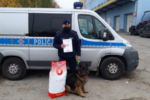 policjant stoi obok Bazyla na tle dużego radiowozu. Obok nich stoi duża karma dla psa. Policjant trzyma w ręku list z podziękowaniami