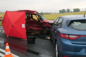 dwa rozbite auta koloru czerwonego i niebieskiego ustawione do siebie na przeciwko stoją przy barierkach. Przy czerwonym aucie rozmieszczony jest czerwony parawan
