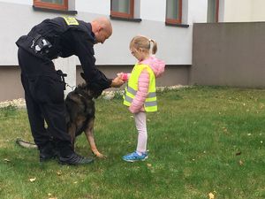 Dziewczynka stoi przy policjanci z psem służbowym i wyciąga w jego stronę rękę, by go pogłaskać
