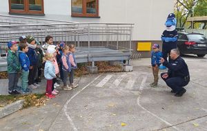 Policjant i policyjny Polfinek rozmawiają z dziećmi na terenie przy przedszkolu.