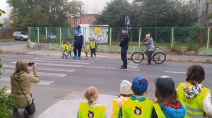 Dzieci wraz z Polfinkiem przechodzą przez przejście dla pieszych. Obok nich stoi policjant, a za nim rowerzysta, który ustępuje pierwszeństwa.