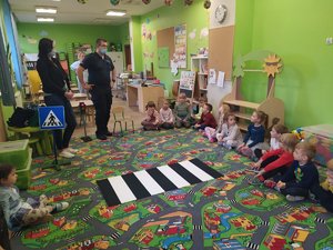 Policjant rozmawia z dziećmi siedzącymi na dywanie w sali przedszkolnej.